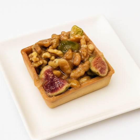 A close-up of a square tart filled with caramelized walnuts, glossy green candied fruits, and sliced figs. The tart sits neatly on a white square plate, against a plain white background.