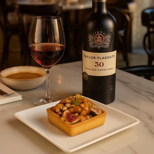 A glass of red wine and a bottle of Taylor Fladgate 30-Year-Old Tawny Port sit on a marble table in a restaurant setting. In front is a small square tart filled with glossy caramelized walnuts, figs, and candied fruits, served on a modern white square plate.