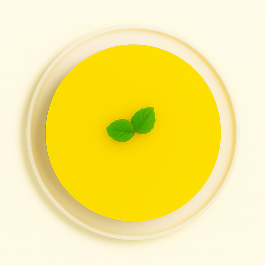 A top-down view of a lemon mousse cake on a white plate. The cake has a smooth, glossy yellow surface with a pair of bright green leaves in the center, set against a plain white background.