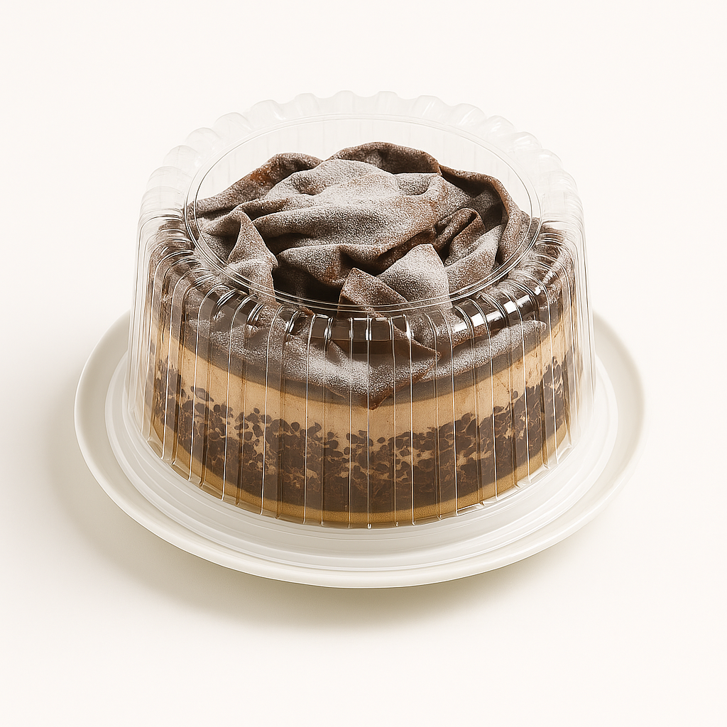 A round chocolate dessert, encased in a clear plastic dome container, sits centered on a white round plate against a plain white background. The dessert features large, folded chocolate curls on top, dusted with powdered sugar, and decorated around the sides with chocolate flakes layered over a creamy filling.